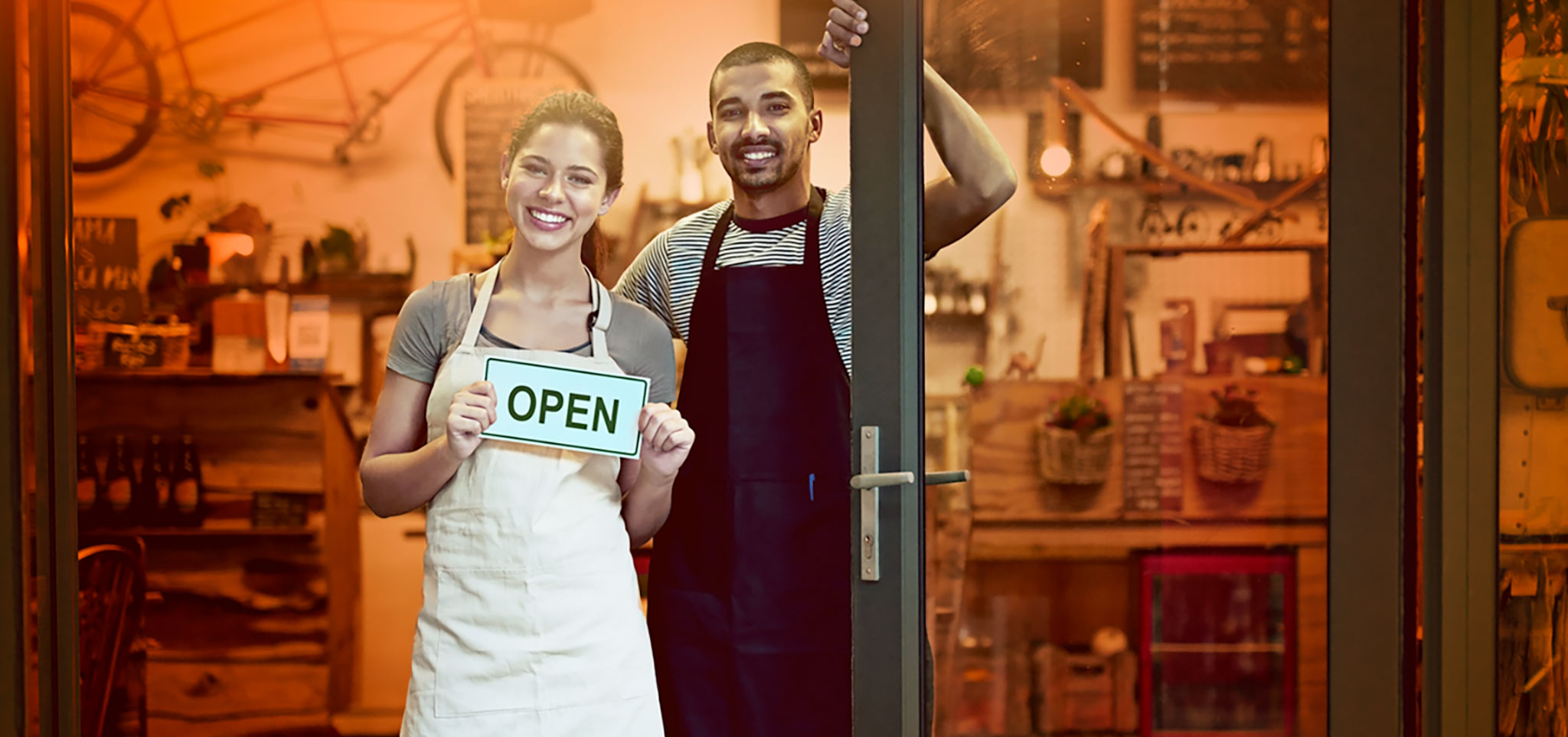 two shop owners in the dorrway of their retail store holding an open sign and smiling.